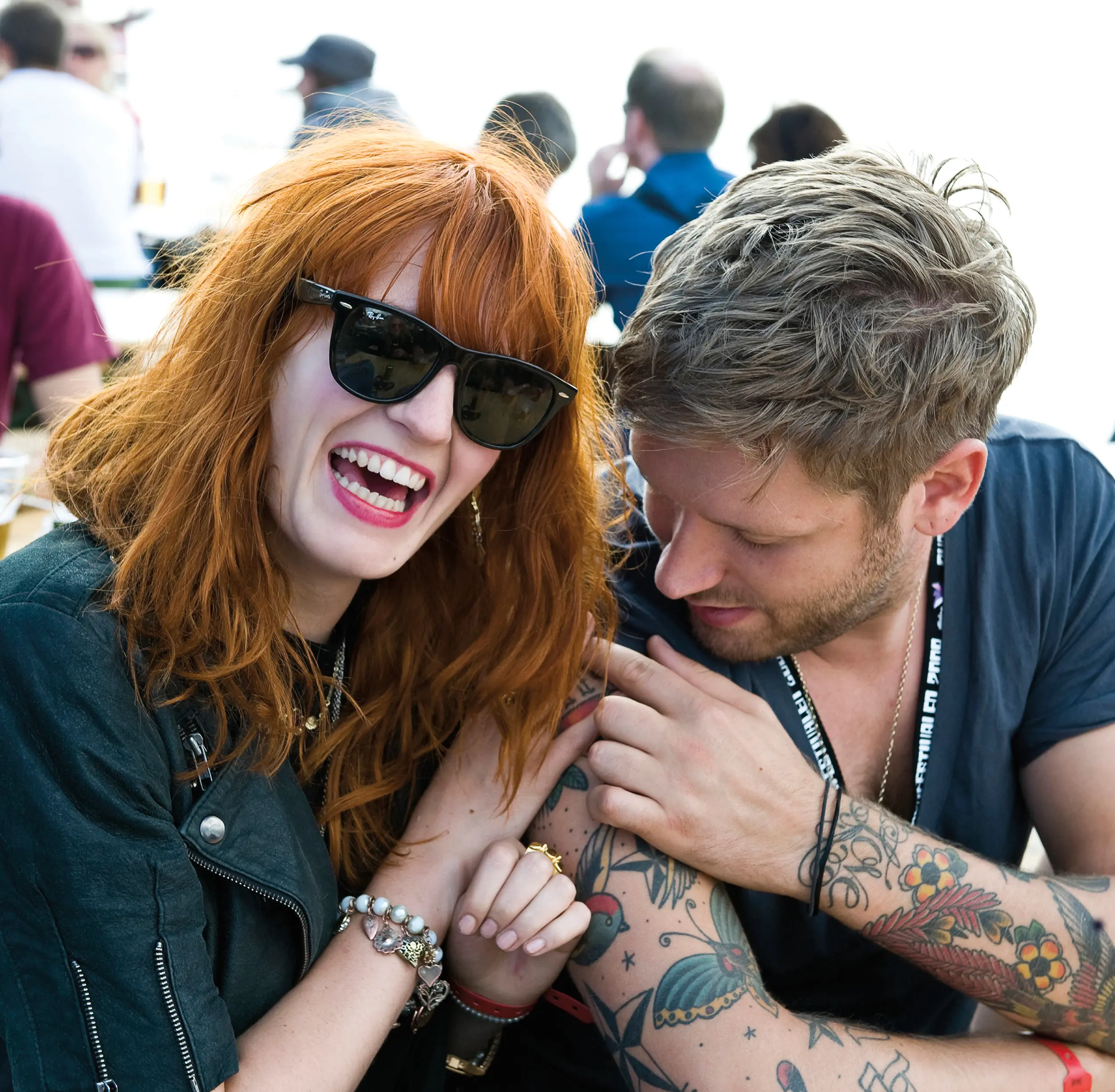 A woman with red hair and Ray-Ban sunglasses laughs while sitting next to a man with tattoos looking at her arm. They are outdoors at a social gathering, with people sitting and talking in the background.